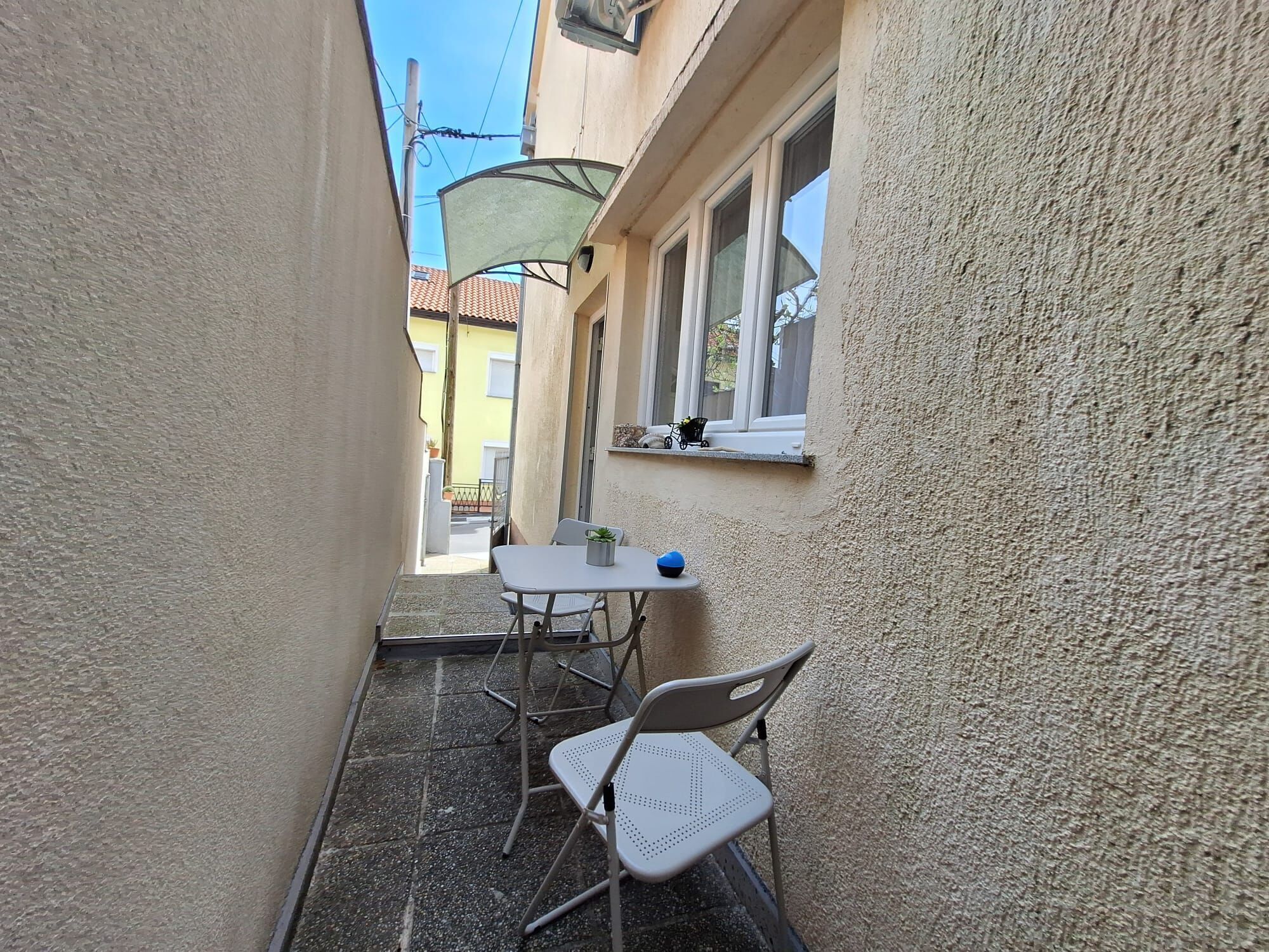Un espacio exterior estrecho con una mesa, sillas, una ventana y un pequeño balcón.