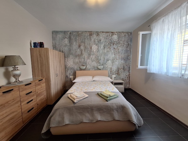 Ein Schlafzimmer mit Bett, Schrank, Nachttisch, Lampe und einem Fenster mit Vorhängen.