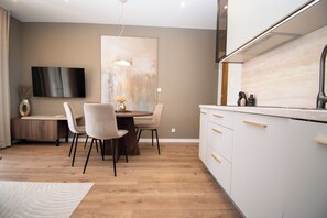 Eine moderne Küche mit weißen Schränken, einem Essbereich mit rundem Tisch und Stühlen, einem an der Wand befestigten Flachbildfernseher und einer Pendelleuchte über dem Tisch.