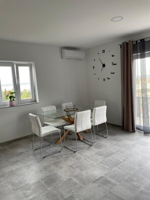 A modern dining area with a glass table and white chairs, a wall clock, and a window with a view. - Apartment Maslina Sea View (Preko)