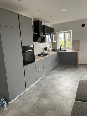 A modern kitchen with grey cabinets, a built-in oven, a stove, and a window providing natural light. - Apartment Maslina Sea View (Preko)