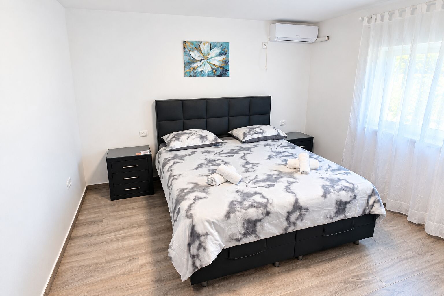 A bedroom with a bed, two bedside tables, a wall-mounted air conditioner, and a window with sheer curtains.