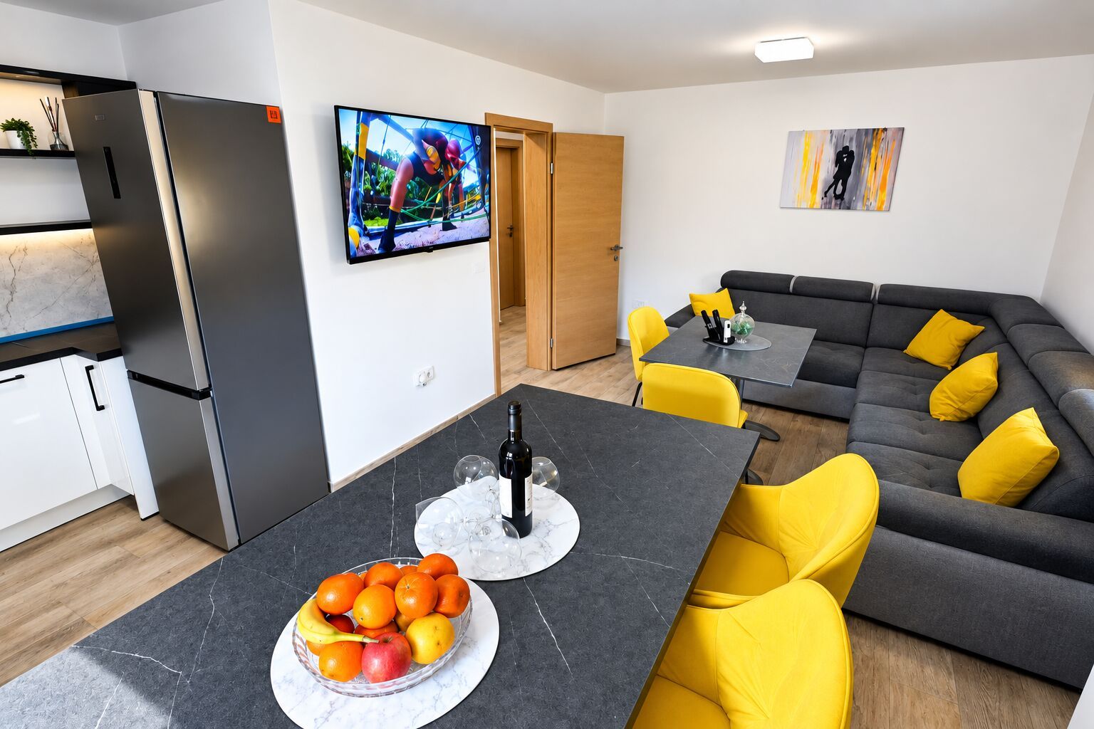 Eine moderne Küche mit integriertem Kühlschrank, einem Essbereich mit schwarzem Tisch und gelben Stühlen sowie ein graues Sofa mit gelben Kissen.