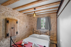 A bedroom with a stone wall, wooden beams, a metal bed frame, a window with curtains, and a hanging light fixture. - Apartment Heritage Stonehouse (Preko)