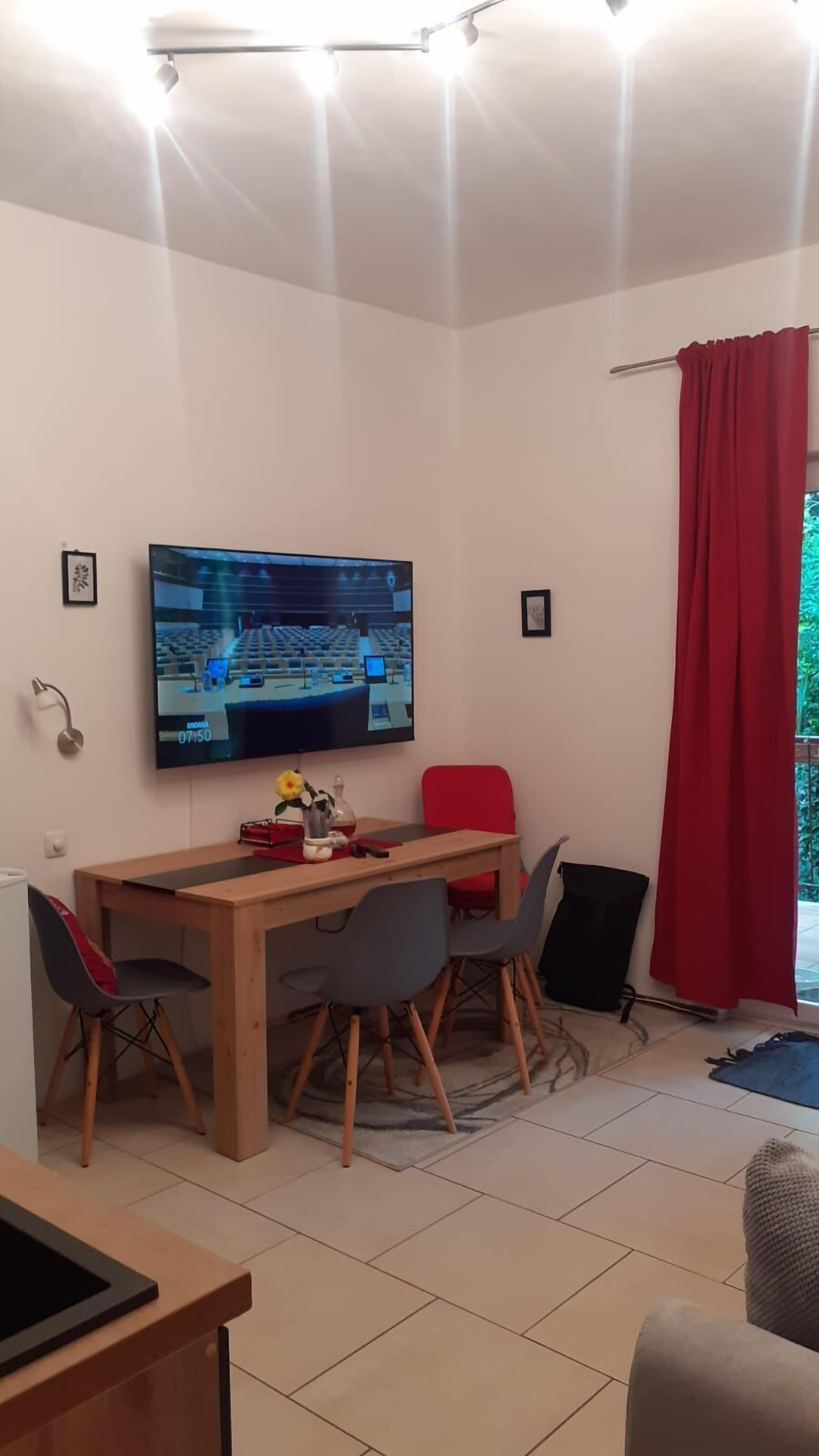Zona pranzo con tavolo in legno, quattro sedie, televisore montato a parete e tenda rossa.