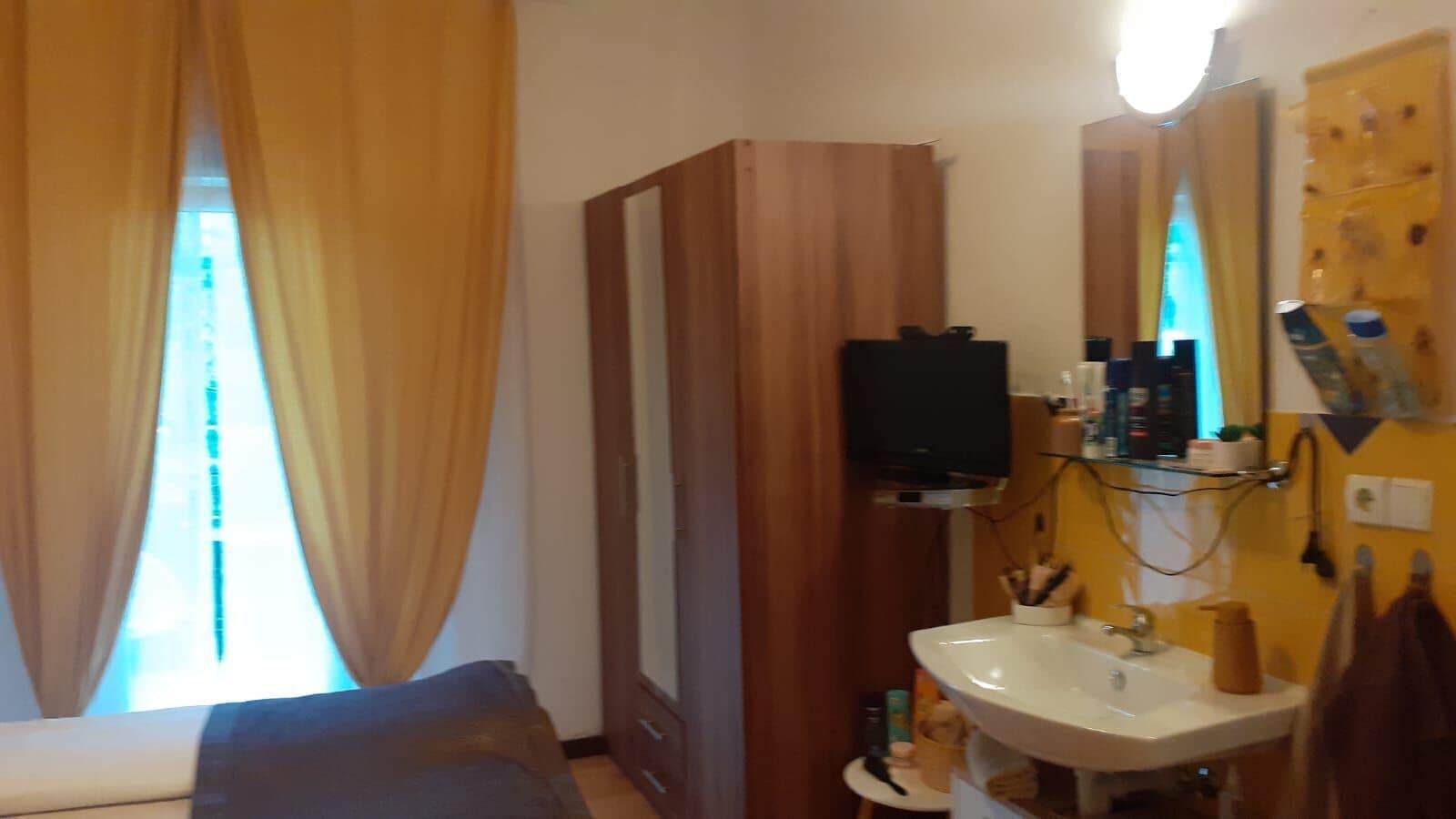 A room with a bed, a television mounted on a wooden panel, a sink with a mirror, and a window with curtains.