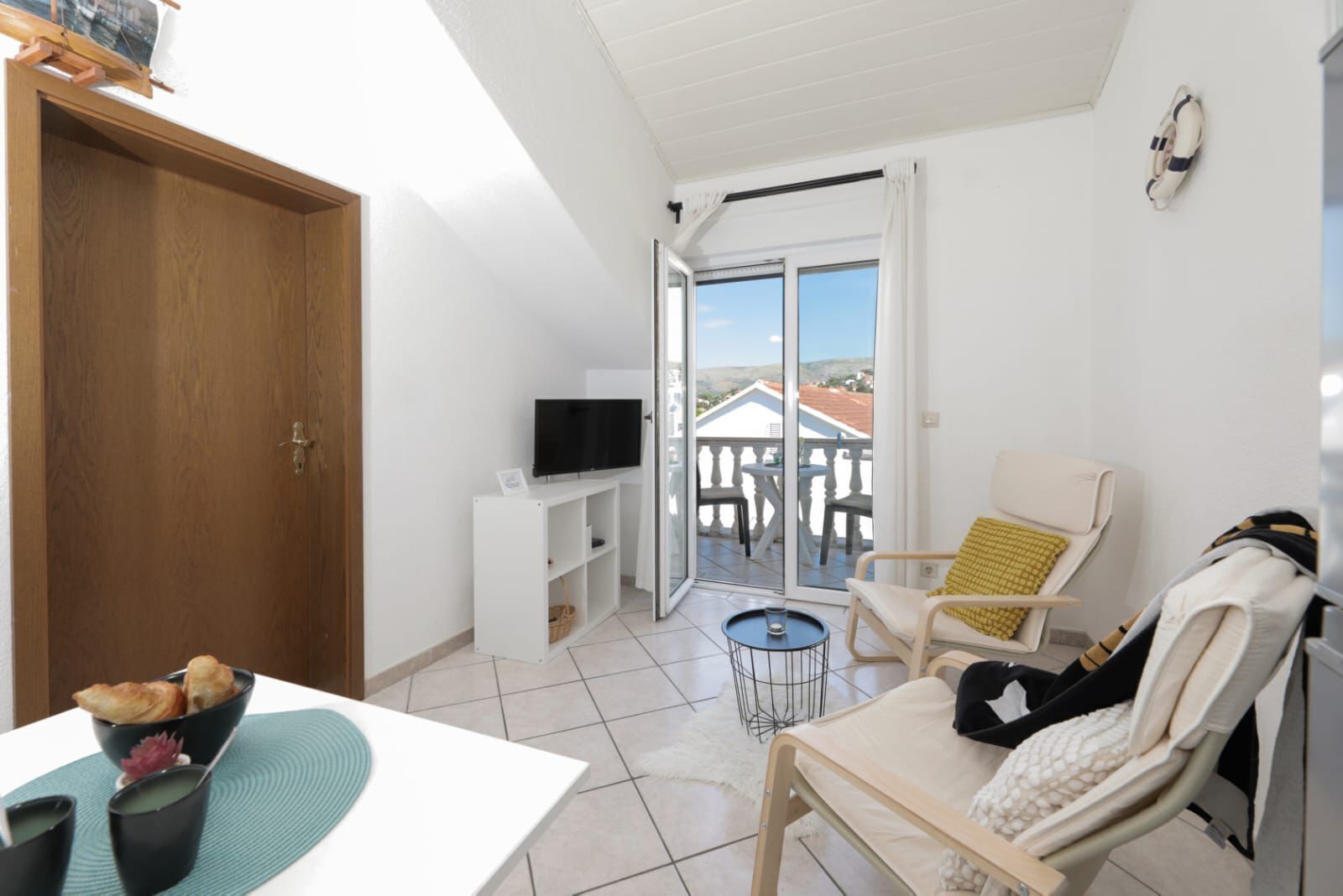 A room with a TV on a white stand, a dining table with a bowl of food, a chair with a cushion, and a balcony with a view.