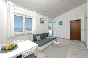 Ein modernes Wohnzimmer mit Sofa, einem Esstisch mit Obst, einem kleinen Beistelltisch und Meerblick durch große Fenster.