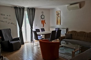 A living room with a sofa, a coffee table, a dining table with chairs, a wall clock, and an air conditioning unit. - House Aida (Sveta Nedelja)