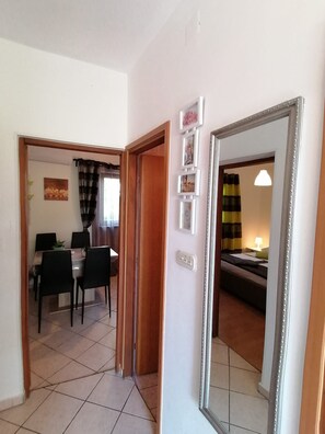 A hallway leading to a dining area with a table and chairs, a bed with a lamp, and a mirror on the wall. - Apartments Andreja (Crikvenica)