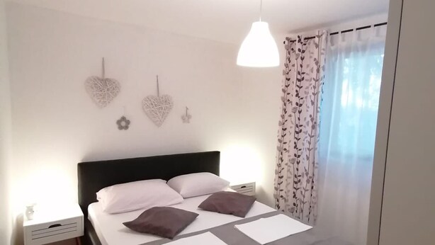 A bedroom with a bed, bedside tables, a hanging light, and a window with curtains. - Apartments Andreja (Crikvenica)