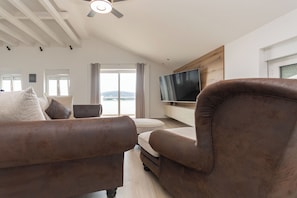 A modern living room with a brown leather sofa, a flat-screen TV mounted on the wall, and a large window with a view of the outdoors. - Apartments Luxury Horizon (Tisno)