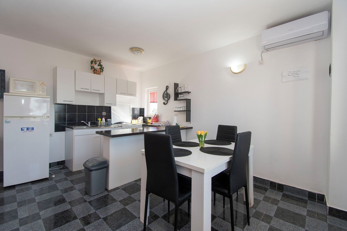 A compact kitchen with white cabinets, a black tiled backsplash, a white refrigerator, and a dining area with a white table and black chairs.