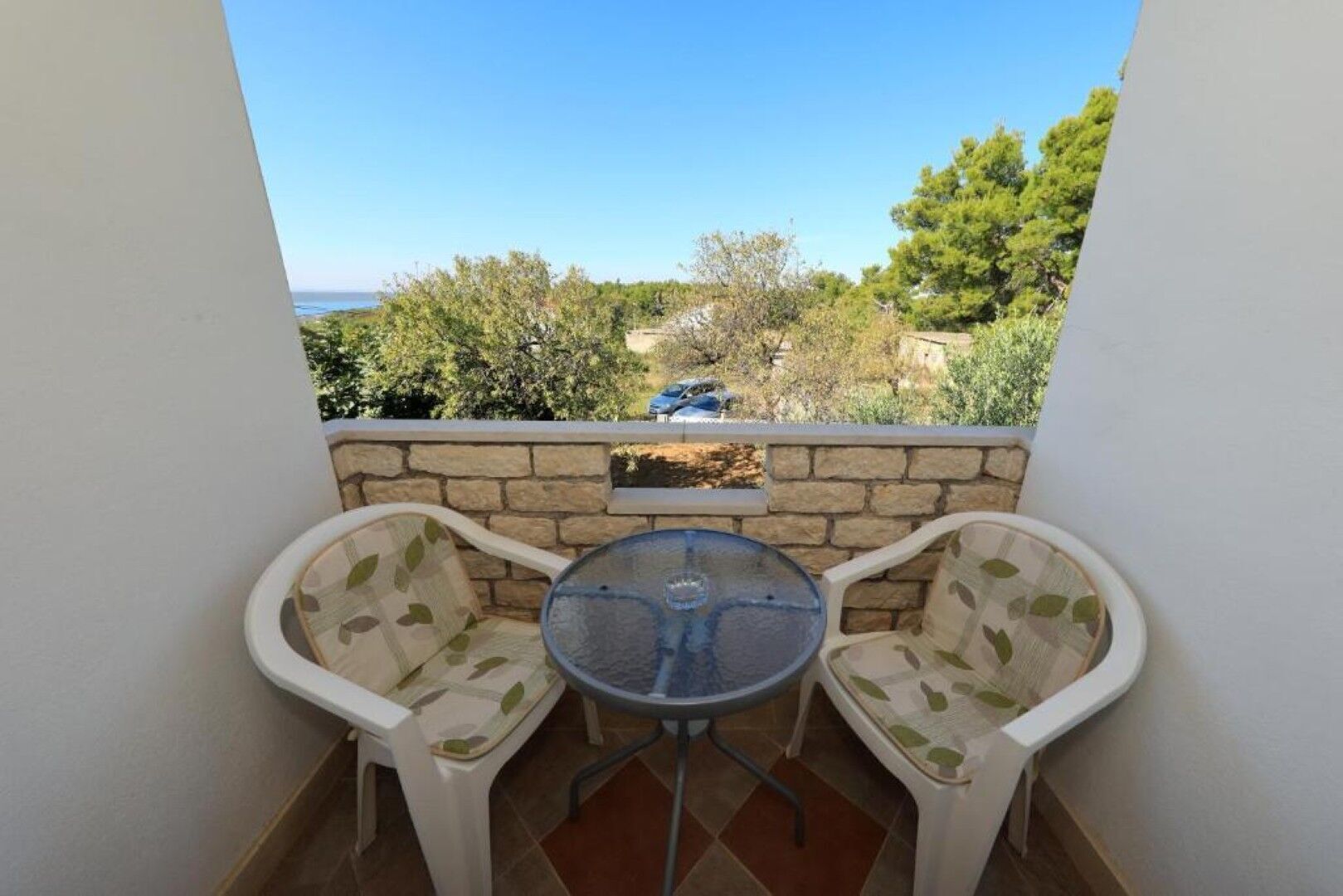 Un petit balcon avec deux chaises blanches et une table, un mur en pierre et une vue sur les arbres et la mer.