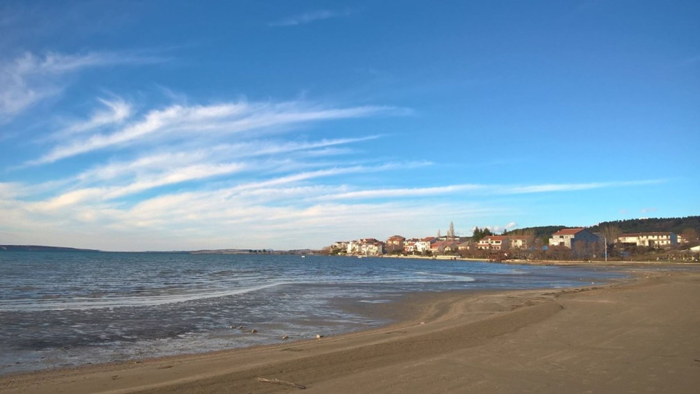 A coastal beach with a clear sky, sandy shore, and buildings in the background.