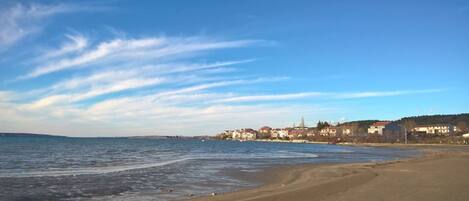 A coastal beach with a clear sky, sandy shore, and buildings in the background.