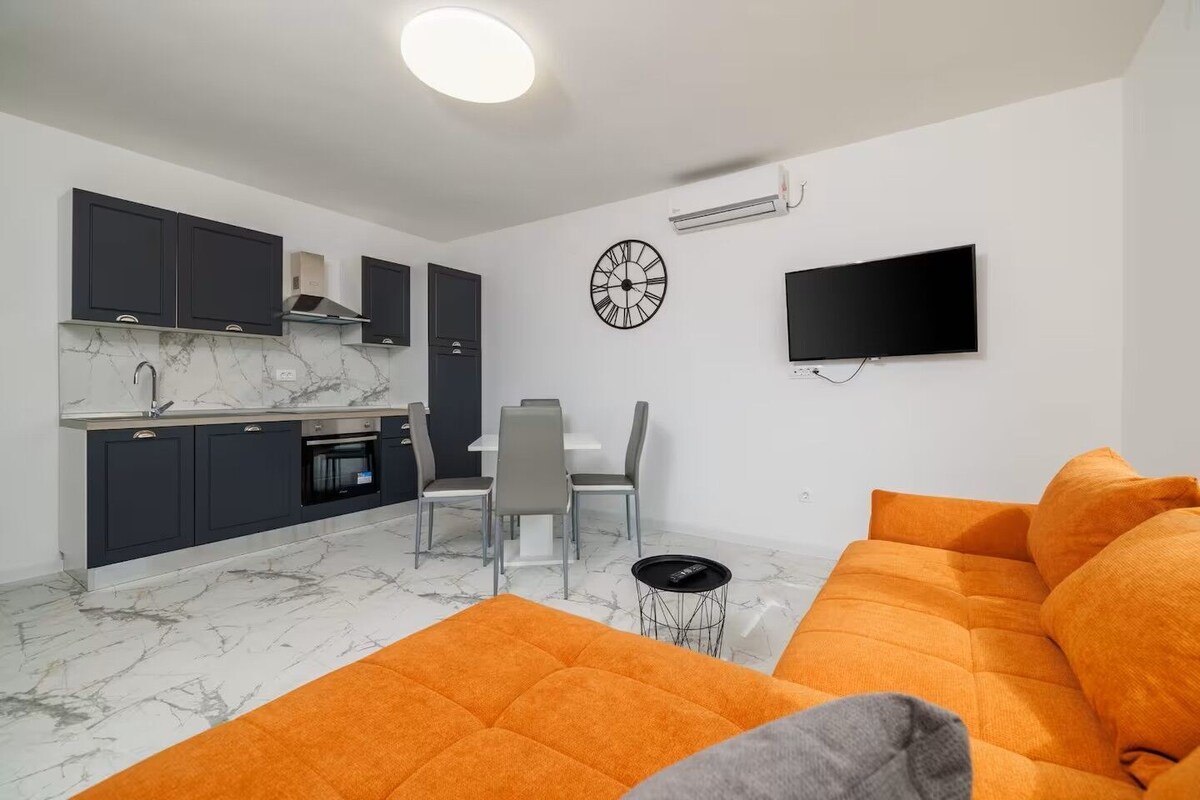 Una sala moderna con un sofá, un comedor con sillas, una cocina con armarios, un televisor y un reloj en la pared.
