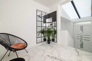 Una habitación con una silla de mimbre negra, una planta en maceta, piso de mármol blanco y un amplio ventanal con vistas.