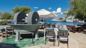 Comedor con mesas y sillas al aire libre, una gran estructura de piedra y vistas al puerto deportivo.