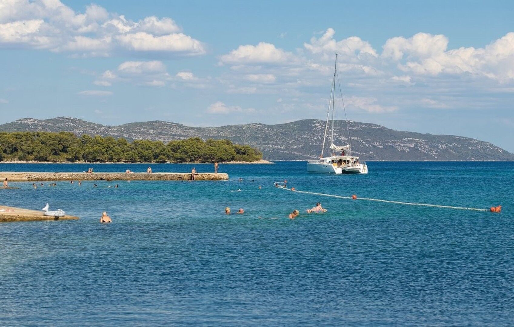 A coastal scene with a sailboat, people swimming, and a rocky shoreline with trees.