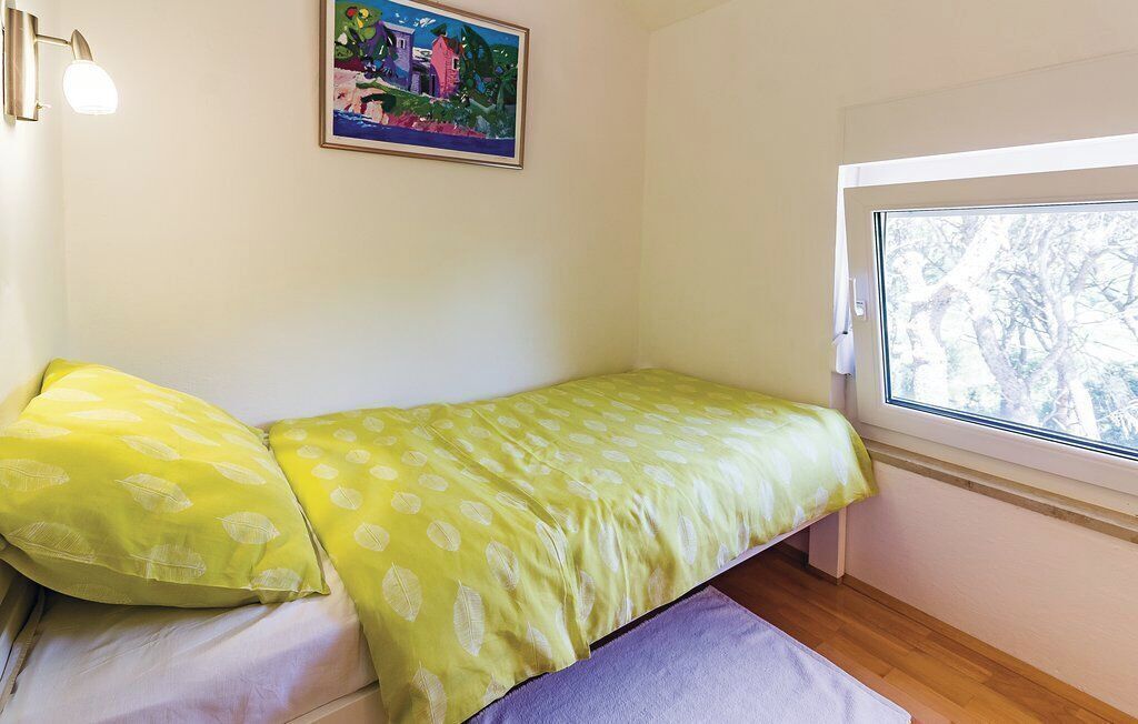 A small room with a bed, a window with a view of trees, a wall-mounted light, and a painting.