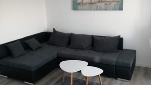 Ein modernes Wohnzimmer mit einem dunkelgrauen Ecksofa, zwei weißen runden Tischen, einem grauen Teppich und einem Gemälde von Segelbooten an der Wand.