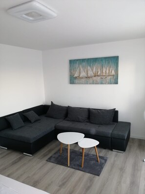 Ein modernes Wohnzimmer mit einem dunkelgrauen Ecksofa, zwei weißen runden Tischen, einem grauen Teppich und einem Gemälde von Segelbooten an der Wand.