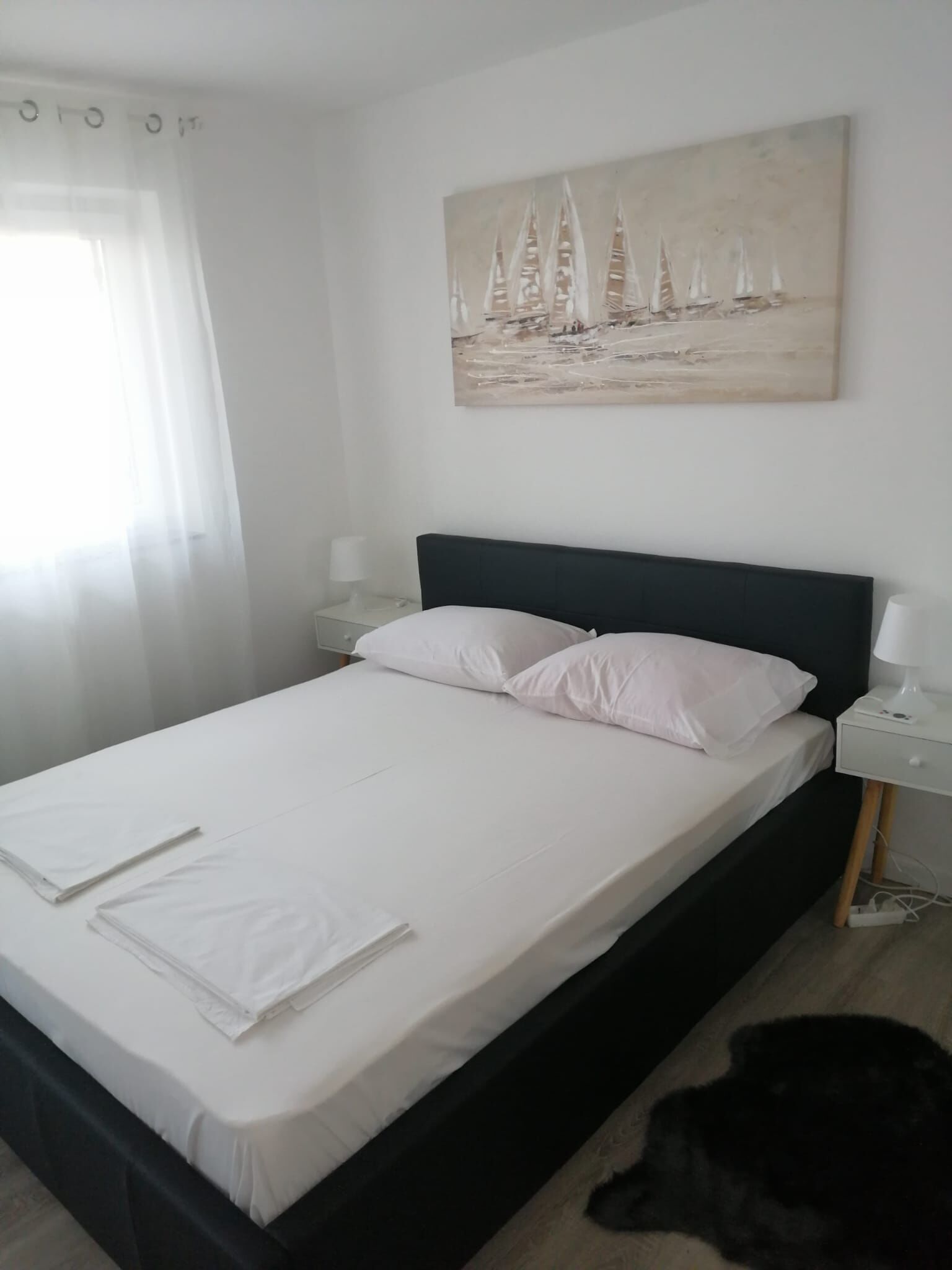 Ein Schlafzimmer mit einem Bett, weißen Laken, einem Segelboot-Gemälde, einem Nachttisch und einer Lampe.