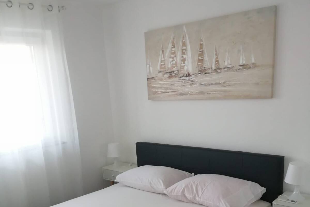 Un dormitorio con cama, ropa de cama blanca, un cuadro de veleros, una mesita de noche y una lámpara.