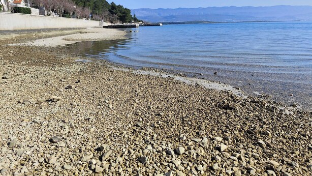Uma praia de seixos com vista clara para o mar e montanhas ao longe.