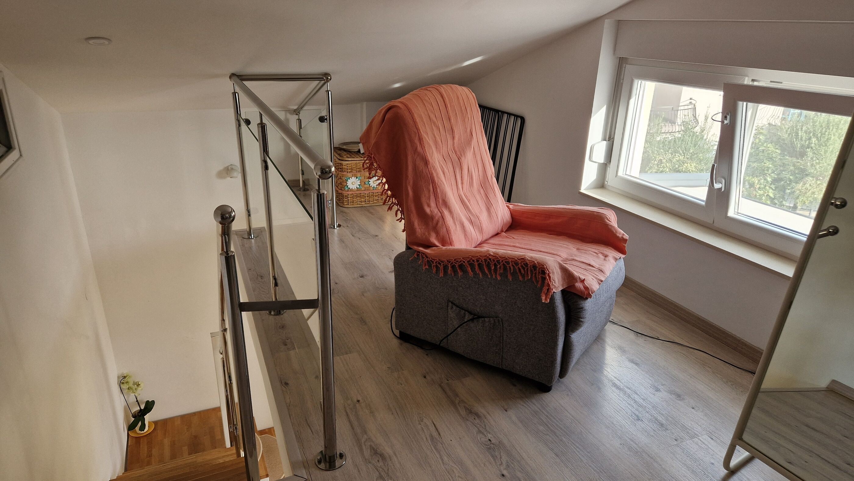 Ein Zimmer mit einem grauen Sessel, der mit einer rosafarbenen Decke bedeckt ist, einer Metallleiter und einem Fenster mit Blick auf Grünflächen.