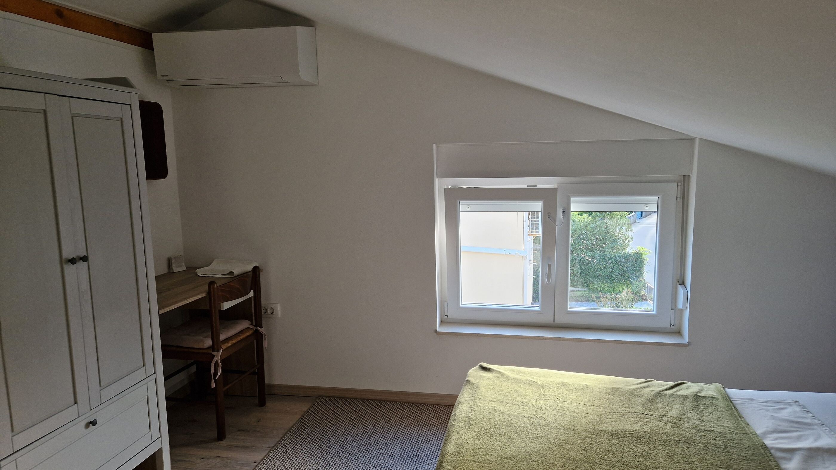 Quarto com teto inclinado, janela com vista para vegetação, cama e guarda-roupa.