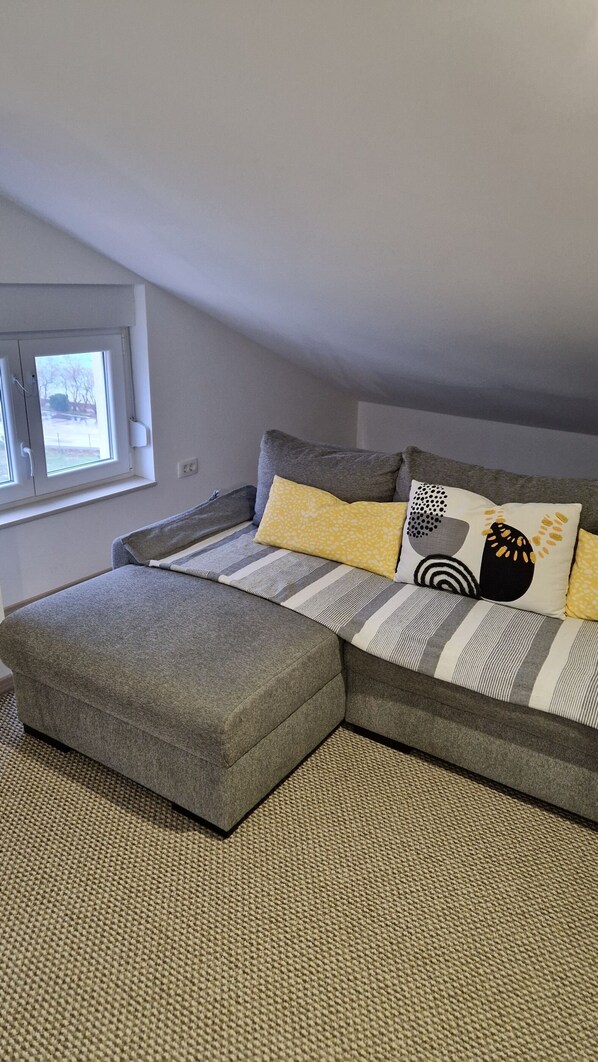 A modern bedroom with a grey sofa bed, a small ottoman, and a window with a view of the outdoors. - Apartment By the sea (Dobrinj)