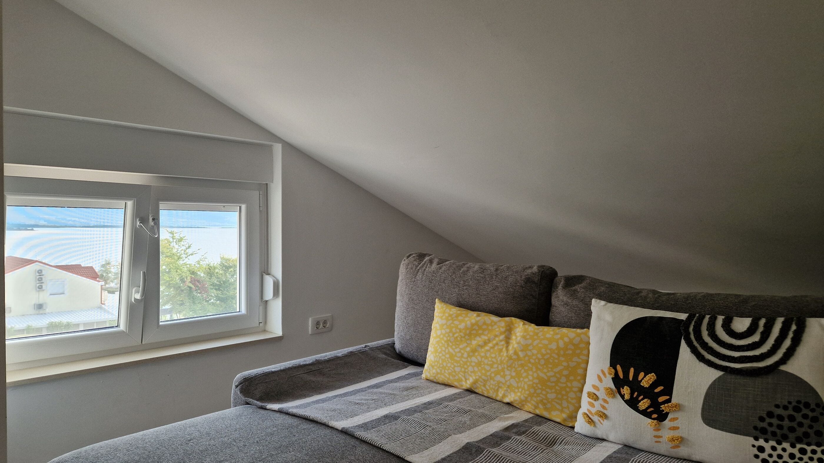Una habitación con techo inclinado, una ventana con vista a edificios, una cama con manta gris y dos almohadas estampadas.