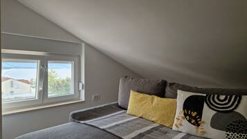 Una habitación con techo inclinado, una ventana con vista a edificios, una cama con manta gris y dos almohadas estampadas.