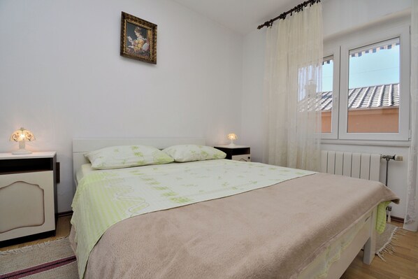 A bedroom with a bed, two bedside tables with lamps, a radiator, and a window with curtains. - Apartment Yolanda (Senj)