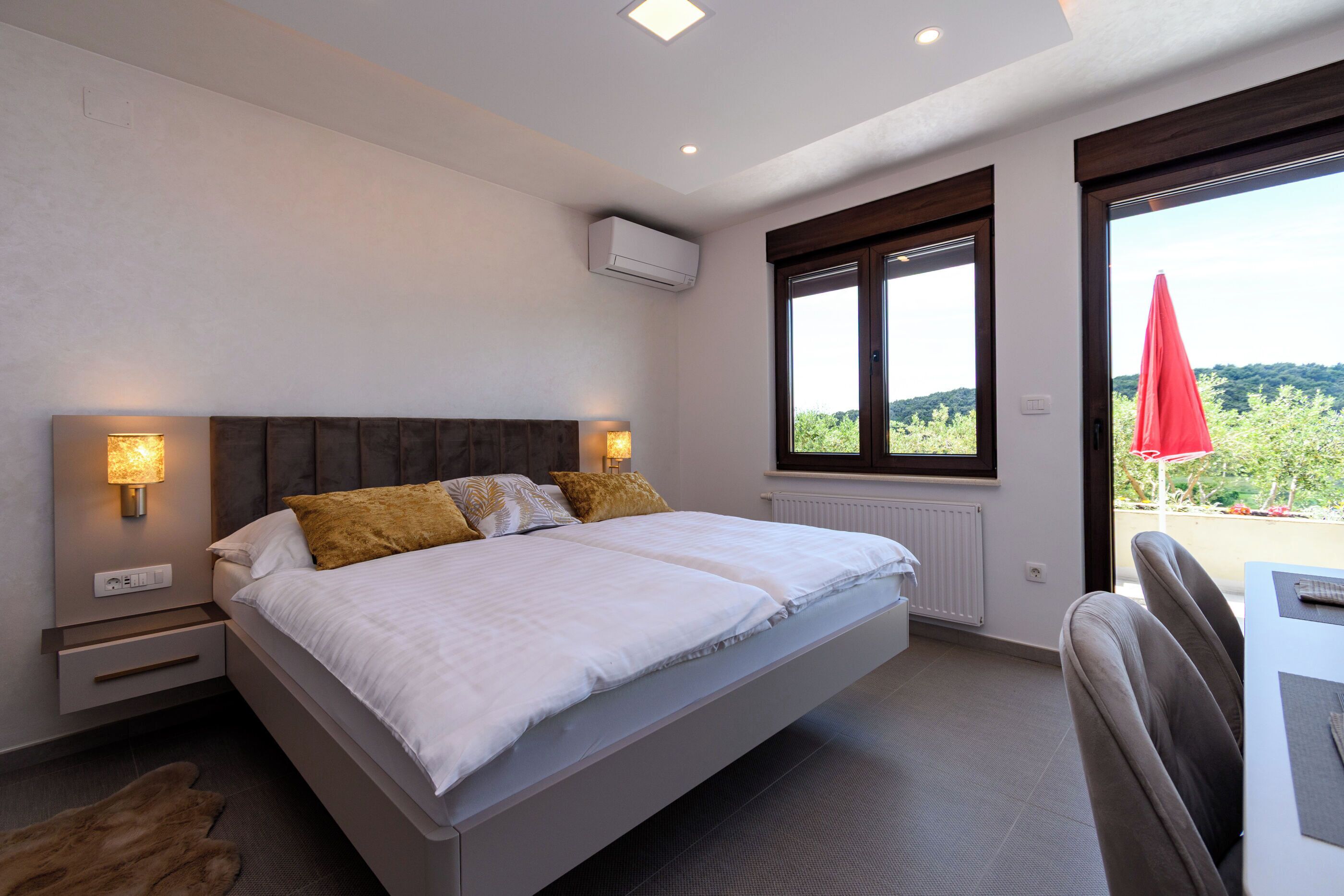 A bedroom with a bed, bedside lamps, a window, and a view of greenery.