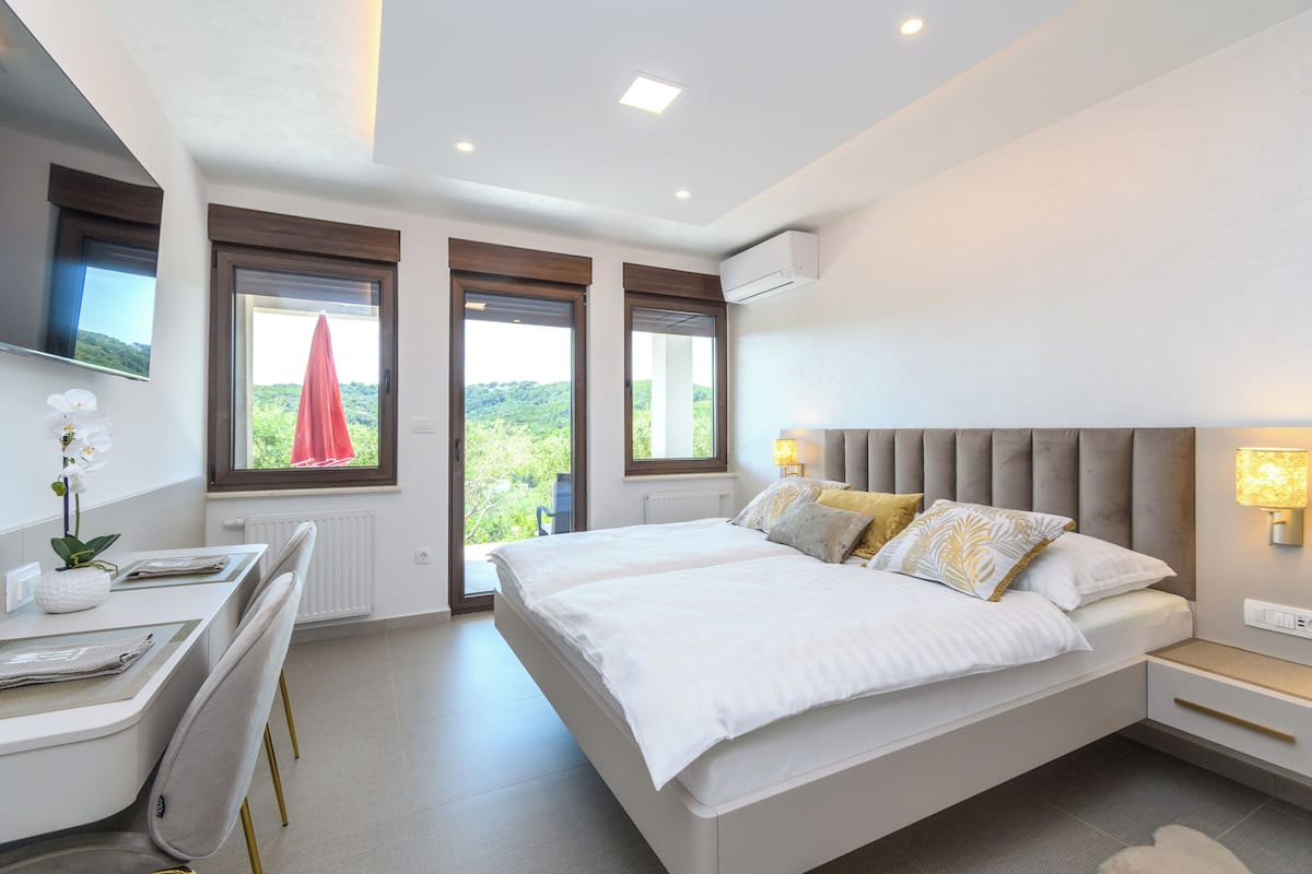 A modern bedroom with a large bed, a desk, and a chair. There are two windows with curtains, an air conditioning unit, and a view of greenery outside.