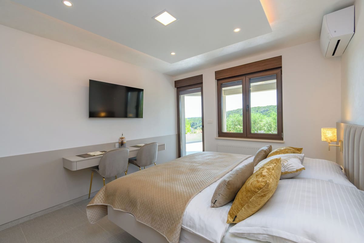 A bedroom with a bed, a desk, a chair, a TV, and a window with a view.