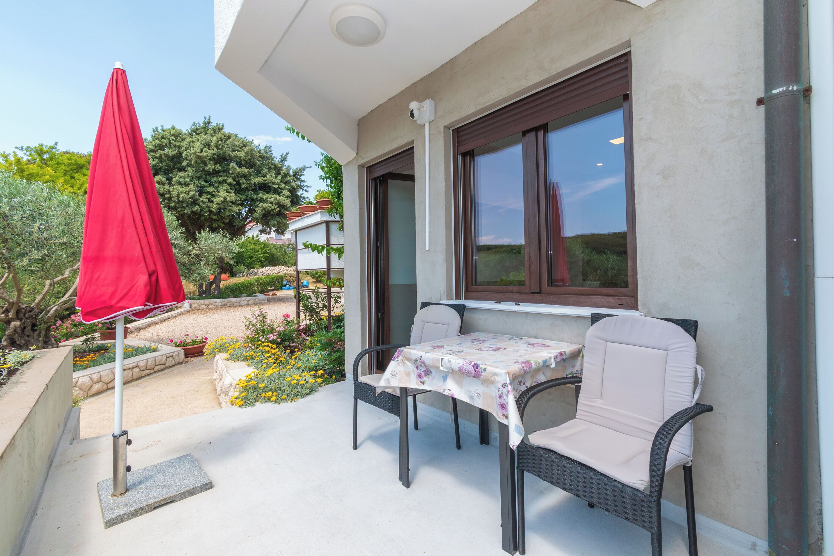 Un patio exterior con mesa y sillas, una sombrilla roja y vista a un jardín.