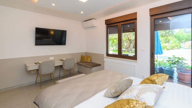 A modern bedroom with a bed, a desk, two chairs, a television, and a window with a view of greenery.