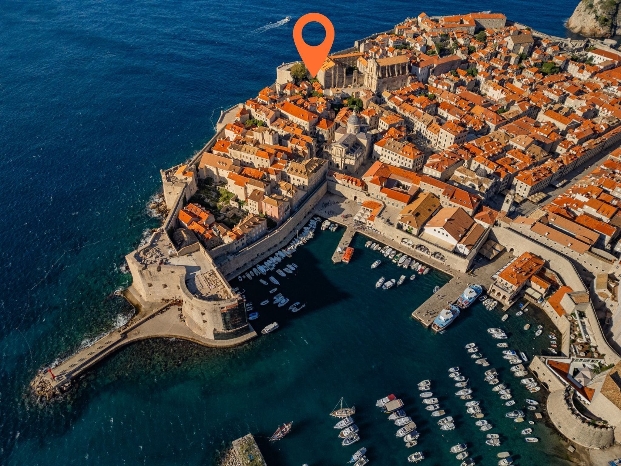 Vista aérea de un pueblo costero con arquitectura histórica, un muro fortificado y un puerto deportivo con numerosos barcos.