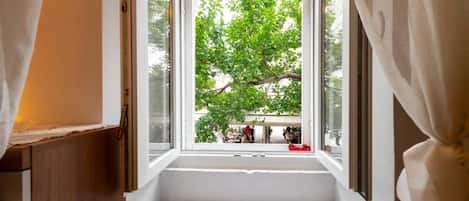 A room with a window, a bed, and a view of a tree and a building outside.