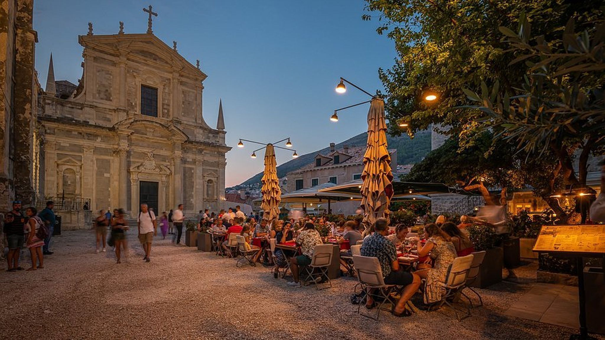Un café en plein air avec des tables et des chaises, un bâtiment historique surmonté d’une croix, et un ciel dégagé.