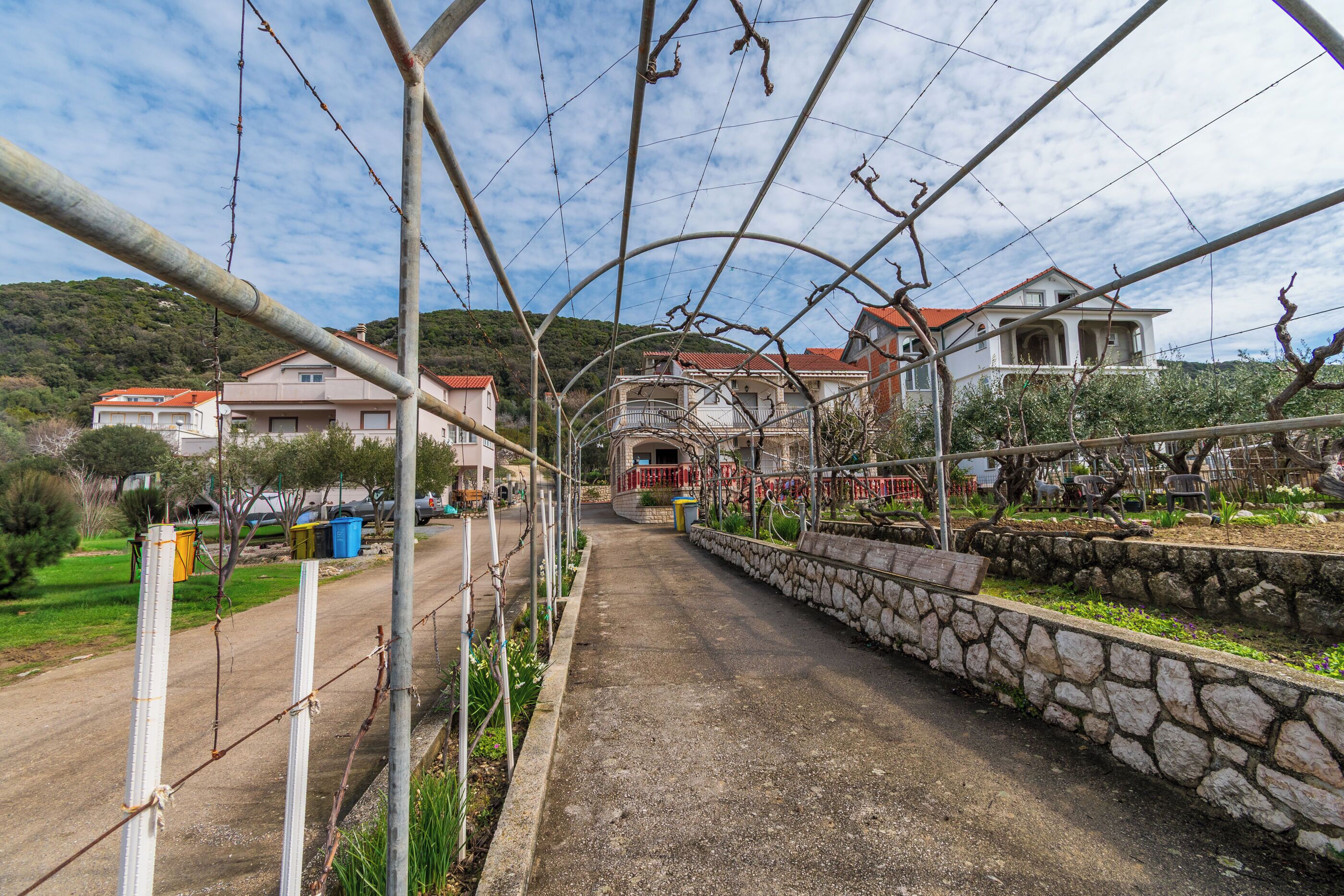 A paved pathway flanked by a stone wall and a barbed wire fence, leading to a building with a red roof and a balcony.