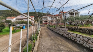 A paved pathway flanked by a stone wall and a barbed wire fence, leading to a building with a red roof and a balcony. - Apartments Maky (Rab)