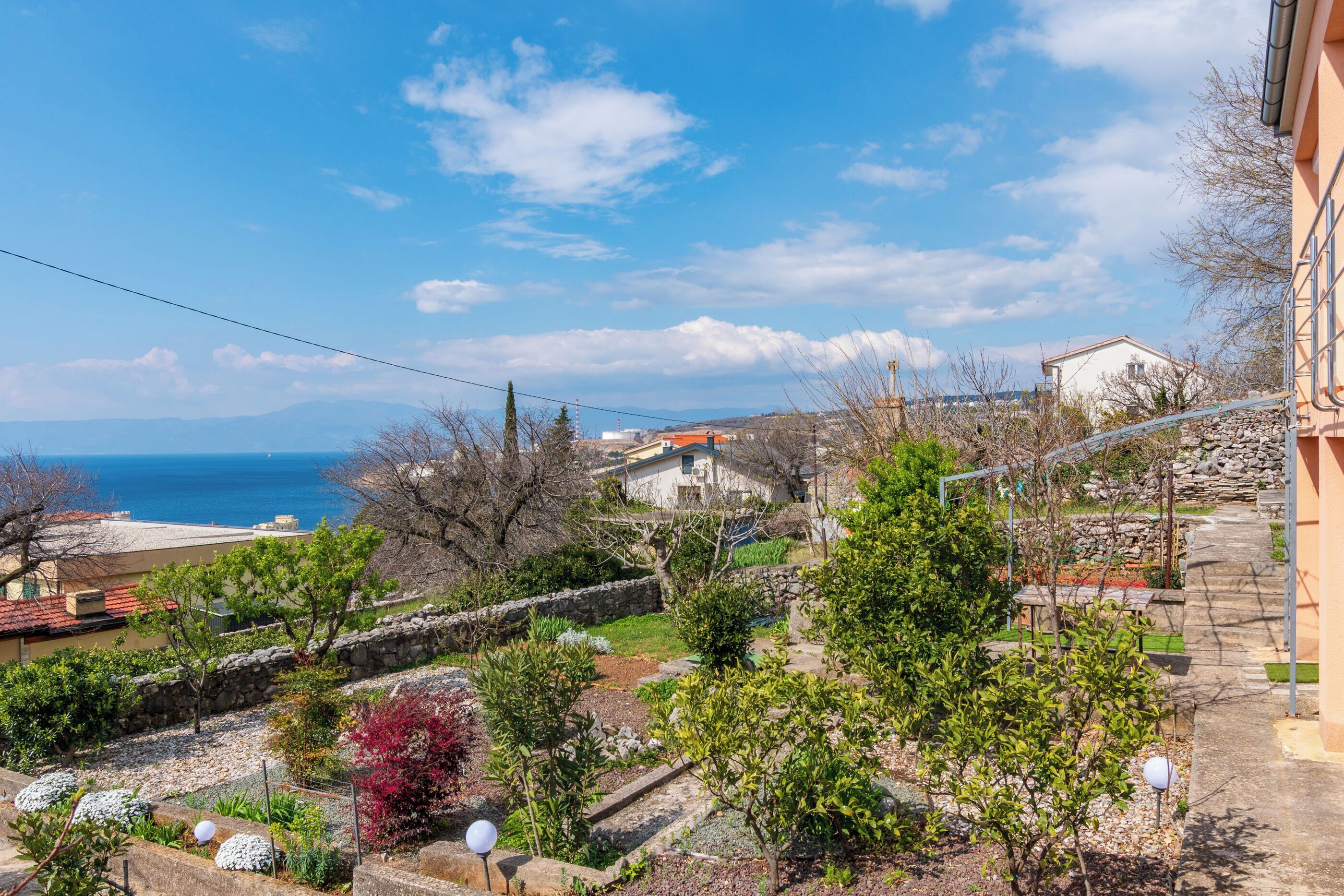 A residential area with a view of the sea, stone walls, and a variety of trees.