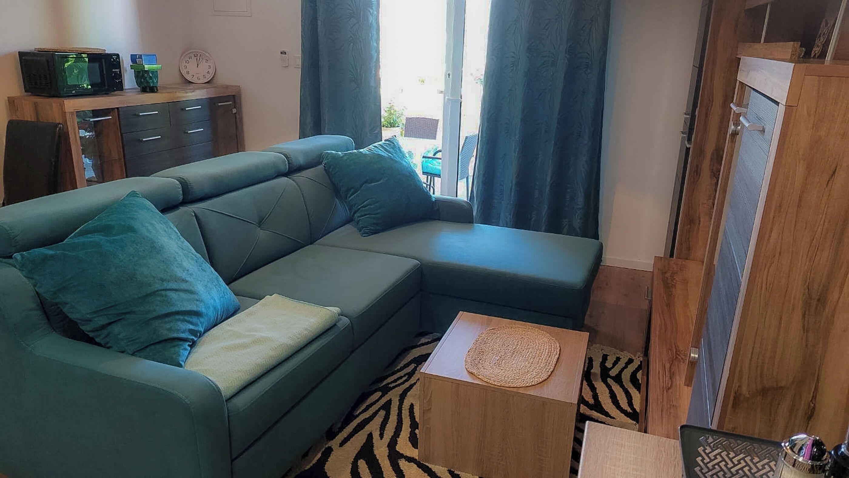 A living room with a sectional sofa, a small wooden side table, a clock, and a view of the outside.