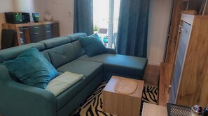 Ein Wohnzimmer mit einer Eckcouch, einem kleinen Holznachtisch, einer Uhr und Blick ins Freie.