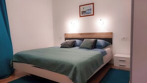 Ein ordentlich bezogenes Bett mit einer grünen Tagesdecke und zwei Kissen, ein hölzernes Kopfteil, ein kleiner Nachttisch mit Schublade und ein gerahmtes Bild an der Wand.
